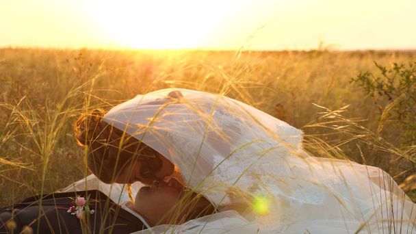 sposa accarezza sposo con erba stelo in faccia. famiglia felice è sdraiato in campo in raggi di bel tramonto. giovane donna bacia il suo amato uomo sulle labbra la sera d'estate sull'erba. famiglia felice
 - Foto, immagini