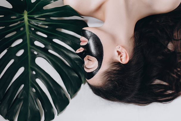 Attractive young girl with eyes closed  with a green leaf on lying bed relaxing with applied a black mask to her face . Woman relaxing with a black facial mask. Concept health and beauty .  - 写真・画像