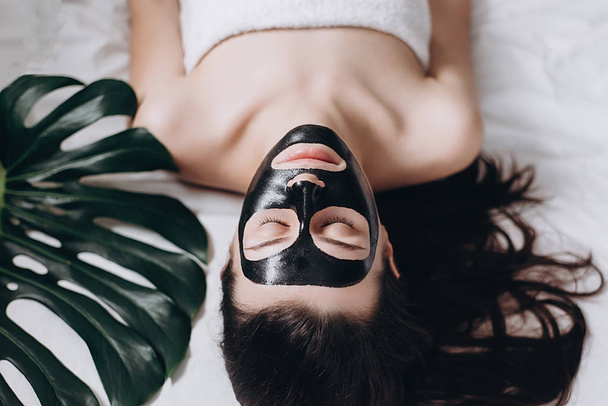 Attractive young girl with eyes closed  with a green leaf on lying bed relaxing with applied a black mask to her face . Woman relaxing with a black facial mask. Concept health and beauty .  - Foto, Imagem