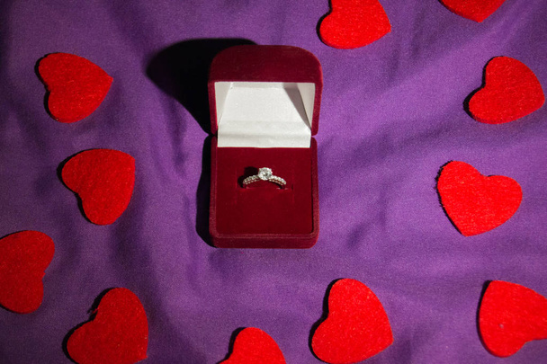 Beautiful wedding ring with precious stone in a red gift box and carved hearts on violet blanked background. Marriage proposal concept. - Photo, image