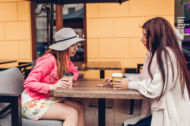 Two women chatting while having coffe in outdoor cafe. Happy friends using phone checking pictures. Girls hang out and having fun - Φωτογραφία, εικόνα