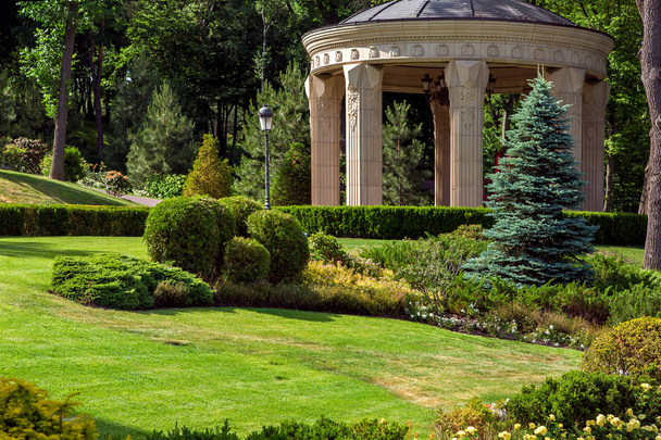 Кам'яна альтанка з колонами в парку з ландшафтним дизайном і зеленими літніми рослинами на передньому плані з газоном і кущами, на фоні листяних дерев
. - Фото, зображення