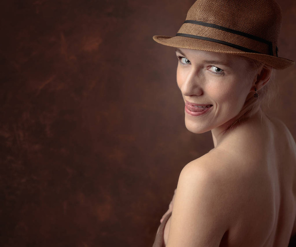 Close-up portret van een mooi midden leeftijd vrouw met blote schouders op een bruine achtergrond. Mooie en slanke vrouw in een hoed toont haar tong. Kopiëren van ruimte. - Foto, afbeelding