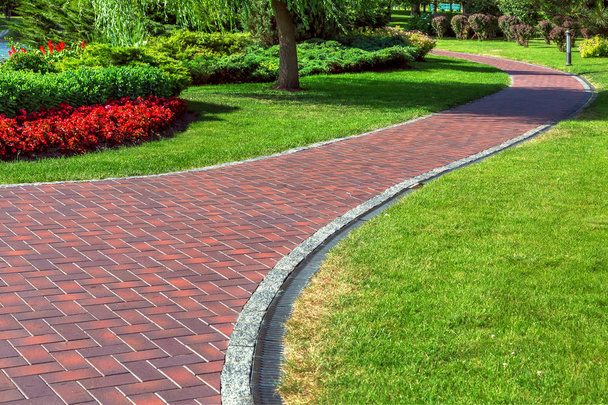 Red blocks footpath with drainage system in a park with green lawns and landscape design with bushes and red flowers. - Фото, зображення