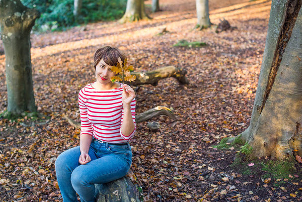 Portrait of young woman sitting and relaxing on the fallen tree in autumnal forest. looking at camera, smiling girl. Autumn outdoor lifestyle. Calm and unity with nature. Selective focus, copy space - Fotó, kép