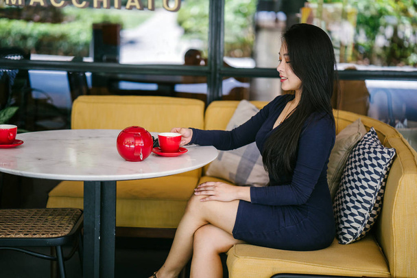 Portrait of a young, attractive, elegant and beautiful Asian woman sitting outdoors in an alfresco cafe and enjoying a hot cup of tea from a red tea set.  - Foto, Bild