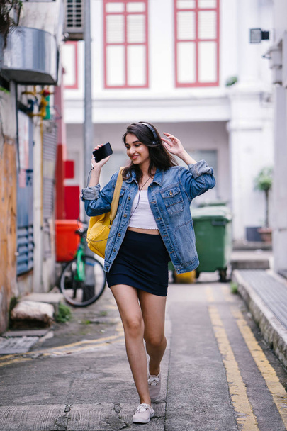Portrait of a young and attractive Indian Asian woman in a denim jacket and yellow backpack enjoying music on her headphones - Foto, Imagem