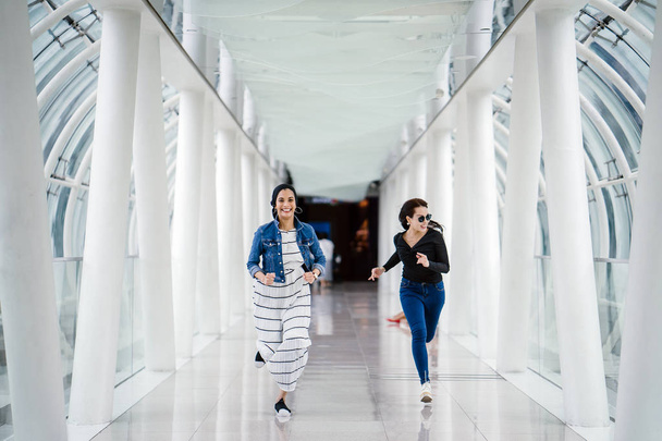 Two diverse friends sprint down a well-lit white futuristic overpass bridge towards the camera. One is a Muslim Middle Eastern woman and the other is Chinese. - Foto, Bild