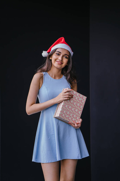 Portrait of a young and beautiful Indian Asian woman in a blue dress and red Santa hat against a black studio background. She is smiling happily as she holds onto a gift for a loved one. - Fotó, kép