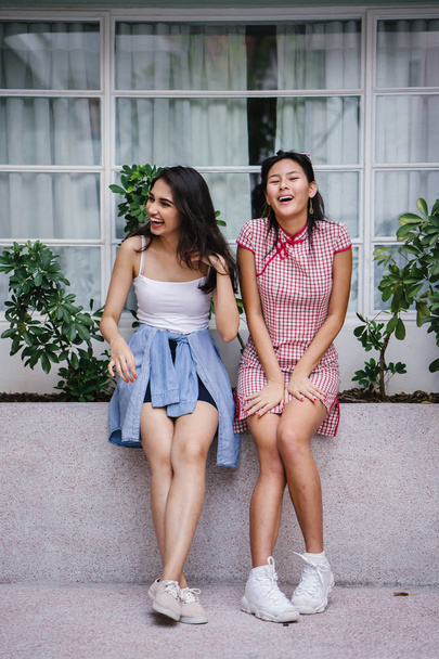 two young women friends of different ethnicity hanging out together. One is a Chinese woman in a cheongsam and the other an Indian woman in a trendy outfit - 写真・画像