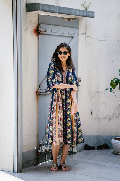 ψηλός, κομψό και όμορφο Ινδική Ασιατική γυναίκα σε ένα καλοκαιρινό φόρεμα και γυαλιά ηλίου χαμόγελα όπως αυτή θέτει σε ένα δρόμο σε ένα στενάκι στη διάρκεια της ημέρας - Φωτογραφία, εικόνα