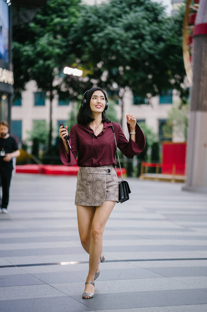 beautiful Korean woman in an elegant blouse and shorts dancing as she walks down a street in the city with her headphones on - Foto, afbeelding