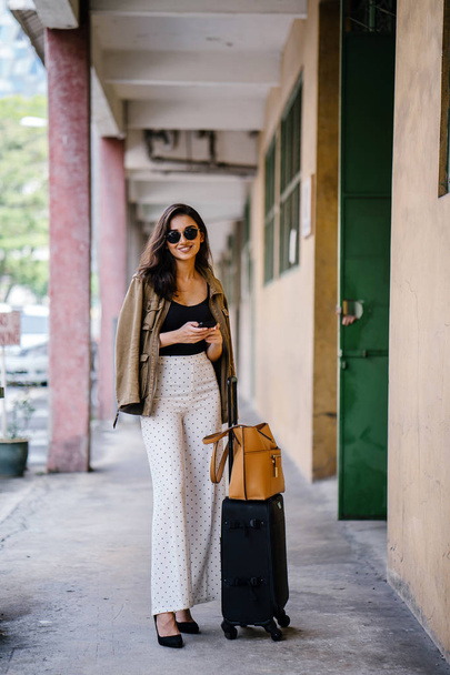 young and attractive Asian Indian woman books a ride through her ride hailing app on her smartphone. She is standing in a walkway with her luggage and is stylishly dressed in work wear and shades - Foto, afbeelding