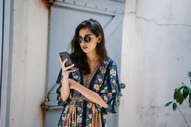 Portrait of a young, attractive and beautiful Indian Asian woman wearing a dress and sunglasses with her smartphone - Foto, Imagen