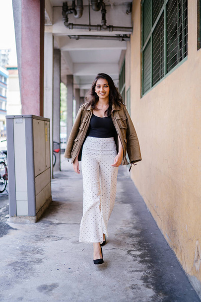 Portrait of a tall, beautiful, slim and elegant Indian Asian woman walking down a walkway in a city in Asia during the day. She is elegantly dressed and has a jacket draped over her shoulders. - 写真・画像