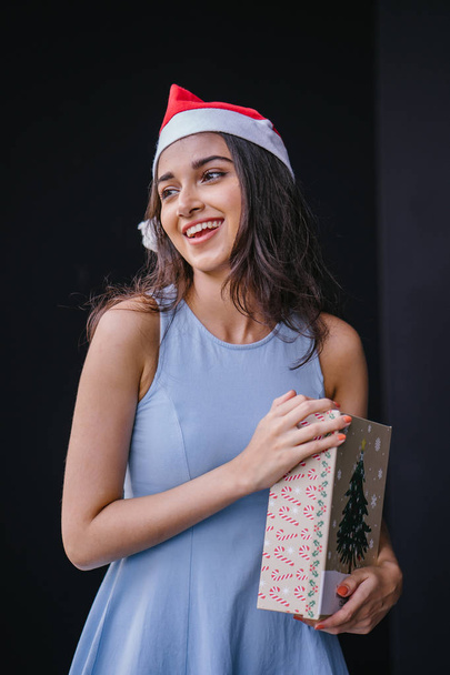 Portrait of a young and beautiful Indian Asian woman in a blue dress and red Santa hat against a black studio background. She is smiling happily as she holds onto a gift for a loved one. - Fotoğraf, Görsel