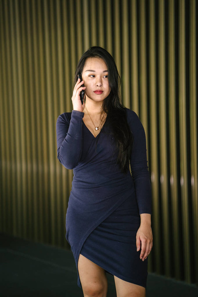 Portrait of a young and beautiful Asian Chinese woman in a classy and elegant blue dress against a modern gold feature wall. She is wearing cool sunglasses as she talks on her smartphone - Zdjęcie, obraz