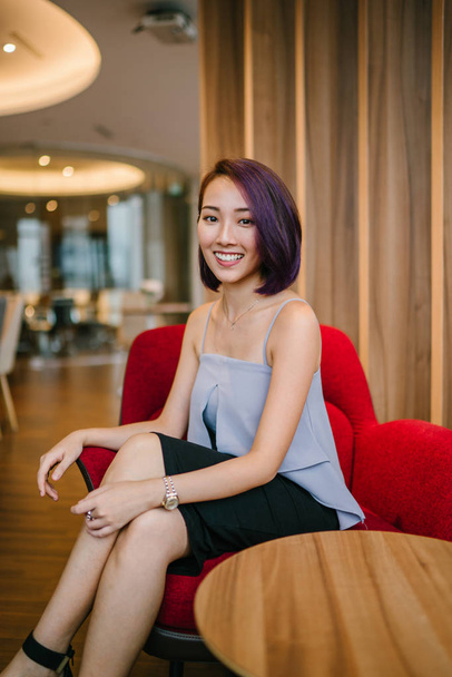 A young, slim and elegant Asian Chinese woman in a professional dress and dyed purple hair sits in a red armchair in her office coworking space. She is smiling candidly. - Foto, Imagem