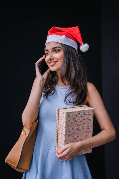 Christmas portrait of a young Indian Asian woman in a red Santa hat smiling as she talks on her smartphone. She is holding on to a present box for a loved one. - Фото, изображение