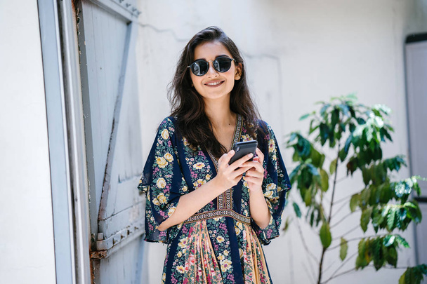 Portrait of a young, attractive and beautiful Indian Asian woman wearing a dress and sunglasses with her smartphone - Foto, imagen