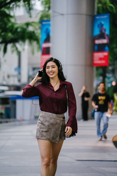 beautiful Korean woman in an elegant blouse and shorts dancing as she walks down a street in the city with her headphones on - Valokuva, kuva