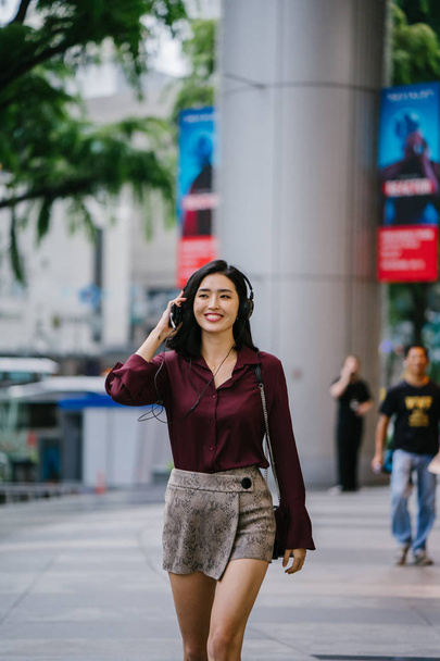 beautiful Korean woman in an elegant blouse and shorts dancing as she walks down a street in the city with her headphones on - Zdjęcie, obraz