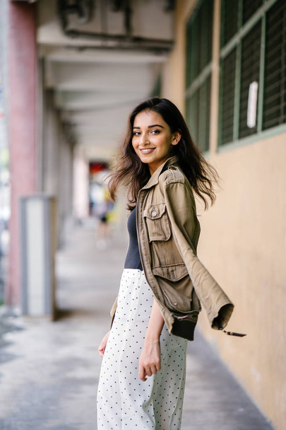 Portrait of a tall, beautiful, slim and elegant Indian Asian woman walking down a walkway in a city in Asia during the day. She is elegantly dressed and has a jacket draped over her shoulders. - Fotó, kép