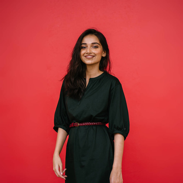Studio portrait of a beautiful, young and attractive Indian Asian woman against a red studio background. She is elegantly dressed in a simple green dress and red belt and is smiling. - Foto, Bild