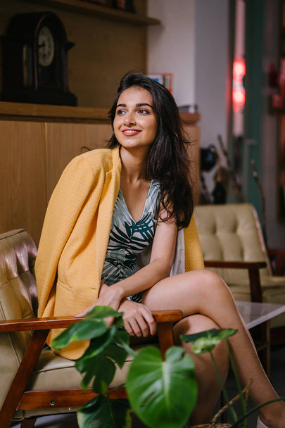  fashionable, beautiful and young Indian Asian woman in a dress and a yellow jacket draped over her shoulders. She is sitting on a chair in a stylish interior - Foto, Imagem