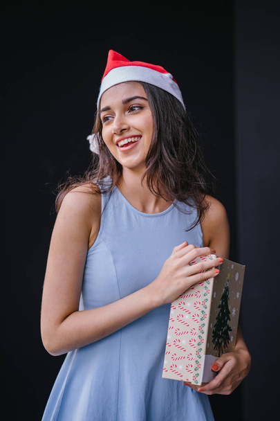 Portrait of a young and beautiful Indian Asian woman in a blue dress and red Santa hat against a black studio background. She is smiling happily as she holds onto a gift for a loved one. - 写真・画像