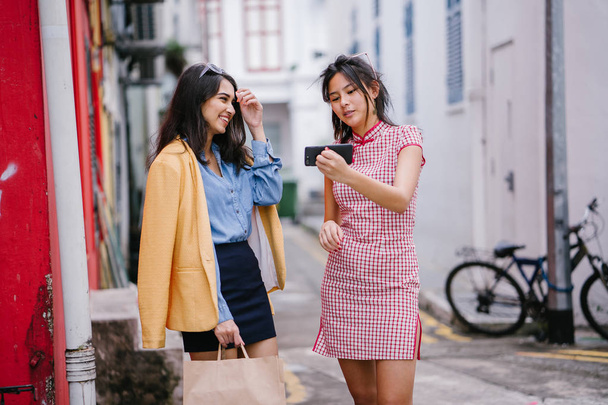 Two young Asian girls have a video call with friends on one of their smartphones. One is Indian, the other Chinese, and they are both fashionably dressed in retro 80's clothing.  - Zdjęcie, obraz