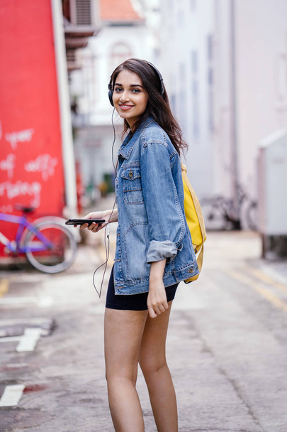 Portrait of a young and attractive Indian Asian woman in a denim jacket and yellow backpack enjoying music on her headphones - Photo, image