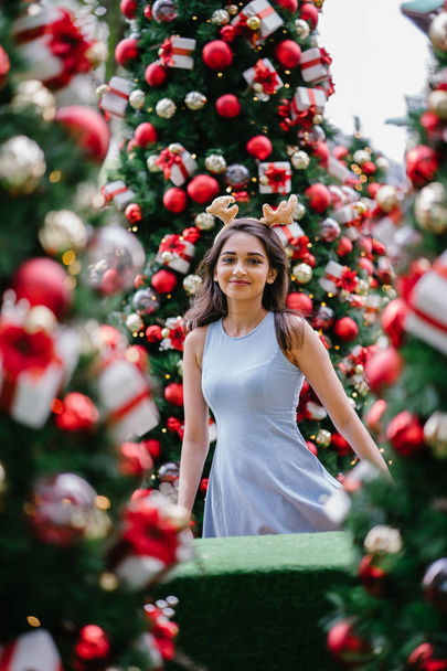 Festive Christmas portrait of a young, beautiful and happy Indian Asian woman smiling as she holds a gift in front of a Christmas tree. - Valokuva, kuva
