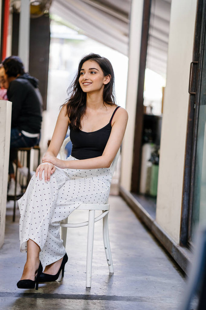 Portrait of a tall, elegant and beautiful Indian Asian girl in a black top and long white pants sitting on a chair in a cafe in the city during the day - Photo, Image