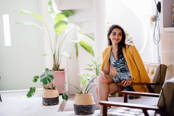  fashionable, beautiful and young Indian Asian woman in a dress and a yellow jacket draped over her shoulders. She is sitting on a chair in a stylish interior - 写真・画像