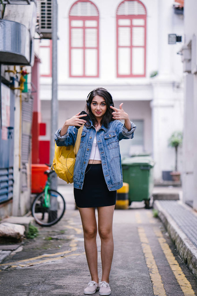 Portrait of a young and attractive Indian Asian woman in a denim jacket and yellow backpack enjoying music on her headphones - 写真・画像