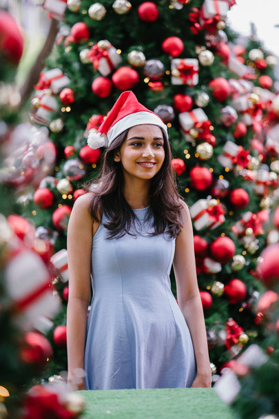 Festive Christmas portrait of a young, beautiful and happy Indian Asian woman smiling as she holds a gift in front of a Christmas tree. - Valokuva, kuva