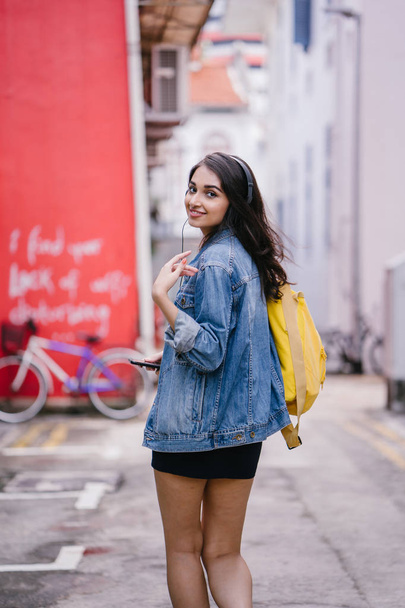 Portrait of a young and attractive Indian Asian woman in a denim jacket and yellow backpack enjoying music on her headphones - Φωτογραφία, εικόνα