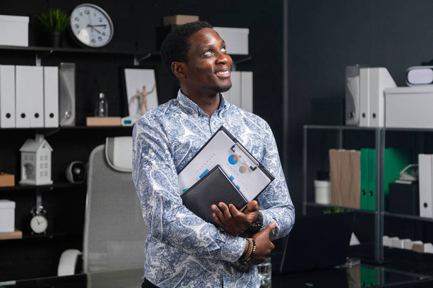 портрет молодого чорного бізнесмена з фінансовими документами в руках, що стоять біля столу в офісі
 - Фото, зображення