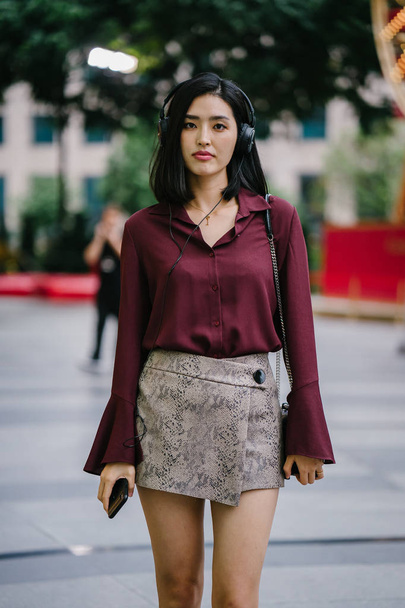 beautiful Korean woman in an elegant blouse and shorts dancing as she walks down a street in the city with her headphones on - Photo, Image