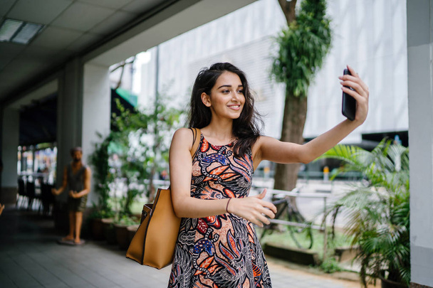 young, beautiful and elegant Punjabi Indian Asian woman takes a selfie of herself with her smartphone on a busy street during the day - Valokuva, kuva