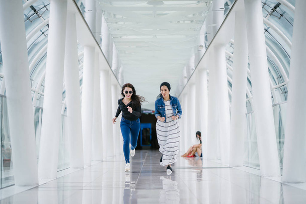 Two diverse friends sprint down a well-lit white futuristic overpass bridge towards the camera. One is a Muslim Middle Eastern woman and the other is Chinese. - Photo, image