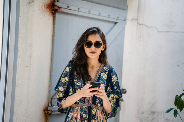 Portrait of a young, attractive and beautiful Indian Asian woman wearing a dress and sunglasses with her smartphone - Foto, afbeelding
