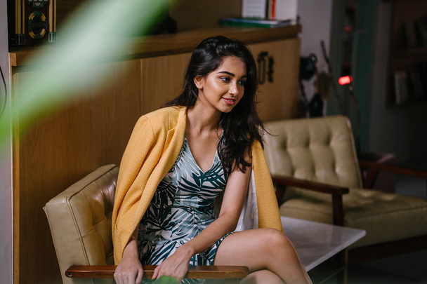  fashionable, beautiful and young Indian Asian woman in a dress and a yellow jacket draped over her shoulders. She is sitting on a chair in a stylish interior - Foto, imagen