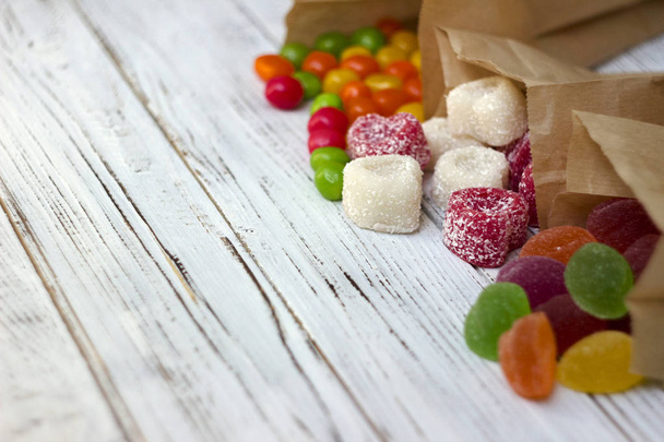 Dulces de colores redondos y mermelada en una bolsa de papel sobre una mesa de madera, dulces brillantes, fondo blanco
 - Foto, Imagen