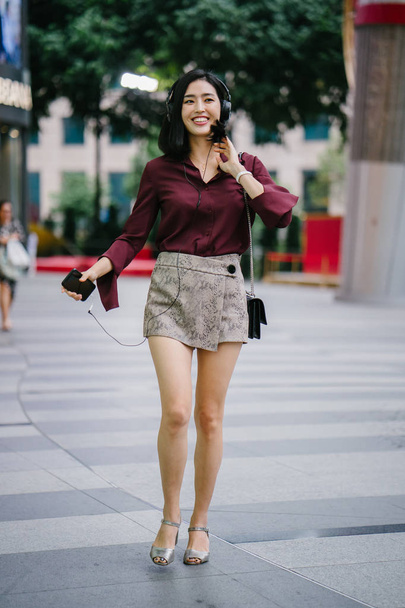 beautiful Korean woman in an elegant blouse and shorts dancing as she walks down a street in the city with her headphones on - Φωτογραφία, εικόνα