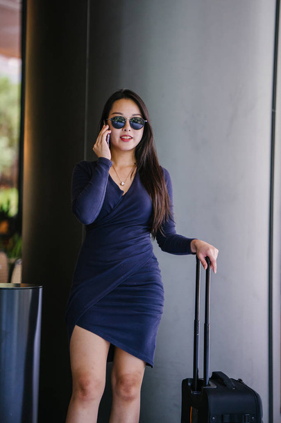 Portrait of an elegant and beautiful young Asian woman waiting on the side of the street for her ride that she booked via a ride-hailing app. She is holding onto a luggage suitcase on wheels.  - Foto, imagen