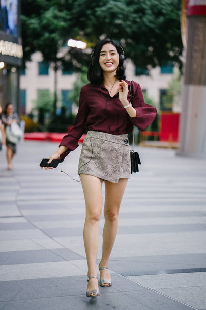 beautiful Korean woman in an elegant blouse and shorts dancing as she walks down a street in the city with her headphones on - Valokuva, kuva