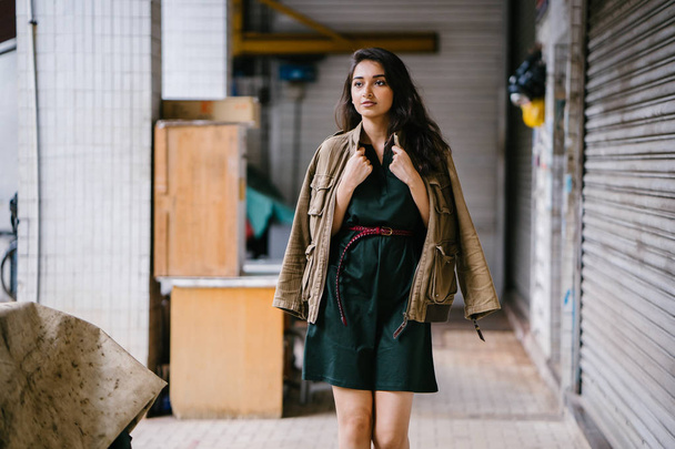 young, tall, slim and elegant Indian Asian woman poses for a portrait. She is wearing comfortable and stylish casual clothing and has a man's jacket draped over her shoulders.  - 写真・画像