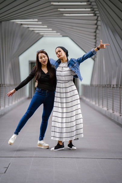 Two friends of different ethnicity on a bridge during the day. One is a Malay Muslim woman and the other is Chinese Asian. They are both young, energetic and attractive. - Фото, изображение